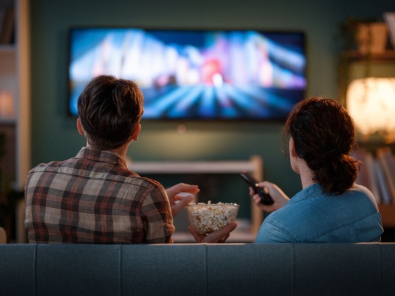 A man and woman watch a movie on a television while sitting on a couch together and sharing popcorn